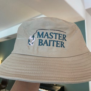 May include: A beige bucket hat with a black and blue embroidered design that says "MASTER BAITER" with a fishing hook and worm.