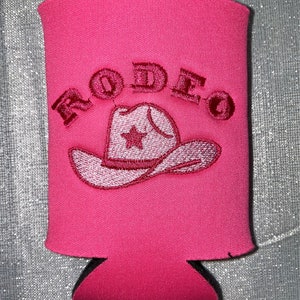 May include: Pink can cooler with a red embroidered cowboy hat and the word "RODEO" above it.
