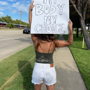 May include: A person holding a sign that says "My Body My Choice". The person is wearing a green tank top with a white graphic that says "1973 Come and Take It" and white denim shorts.
