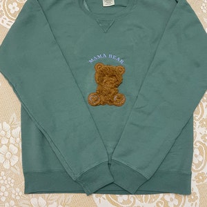 May include: A teal green sweatshirt with a brown teddy bear applique and the text "Mama Bear" embroidered in blue.