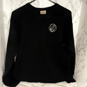 May include: Black crewneck sweatshirt with a white Steal Your Face logo on the chest. The sweatshirt has long sleeves and a ribbed neckline. The Comfort Colors tag is visible at the back of the neck.