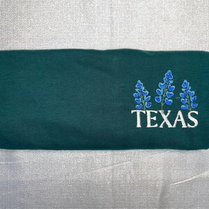 May include: A folded, dark green sweatshirt featuring an embroidered design of bluebonnet flowers and the word "TEXAS" in white. The sweatshirt is made of a soft material and is folded neatly.