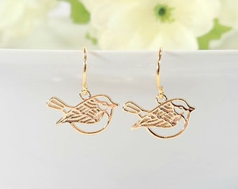 May include: Gold-toned bird earrings with delicate cutouts. The earrings are hanging from a white bowl.