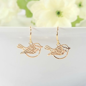 May include: Gold-toned bird earrings with delicate cutouts. The earrings are hanging from a white bowl.