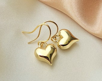 May include: A pair of gold-colored heart-shaped earrings. The earrings have a smooth, polished finish and are suspended from delicate ear wires. The background is a soft, neutral fabric.