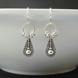 May include: Silver dangle earrings with a teardrop-shaped charm featuring a beaded design and a small round bead at the bottom. The earrings have a hoop at the top with multiple small loops.