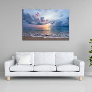 May include: A photograph of a beach scene with a cloudy sky and a sunset over the ocean. The water is a light blue color and the sand is a light brown color.