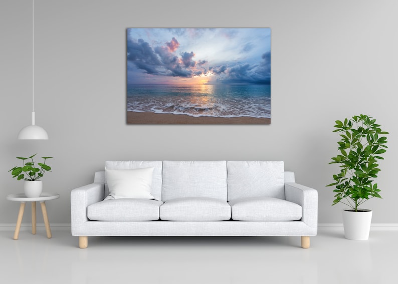 May include: A photograph of a beach scene with a cloudy sky and a sunset over the ocean. The water is a light blue color and the sand is a light brown color.