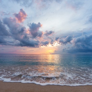 May include: A scenic view of a sandy beach with foamy waves lapping at the shore. The sky is a mix of blue, pink, and gray clouds with a bright sun shining through.