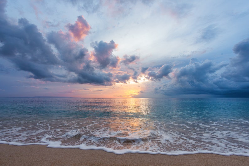 May include: A scenic view of a sandy beach with foamy waves lapping at the shore. The sky is a mix of blue, pink, and gray clouds with a bright sun shining through.