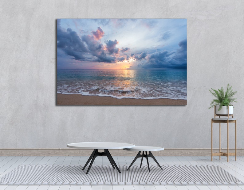 May include: A photograph of a beach scene with a cloudy sky and a sunset over the ocean. The waves are crashing on the shore and the sand is white.