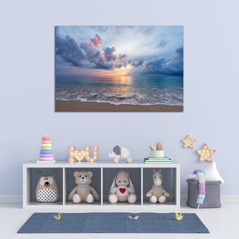 May include: A white shelf unit with four compartments, each containing a different stuffed animal. The top shelf has a wooden elephant toy and a wooden sign that says "I love U". The shelf is against a light blue wall with a canvas print of a beach scene with a sunset and clouds.