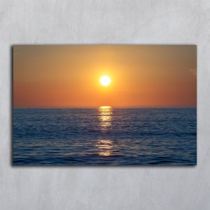 Könnte beinhalten: Ein rechteckiger Wandkunstdruck, der einen leuchtenden Sonnenuntergang über einem ruhigen Ozean zeigt. Die hellgelbe Sonne steht tief am Horizont und wirft eine schimmernde goldene Reflexion auf das tiefblaue Wasser unter einem orangefarbenen Himmel.