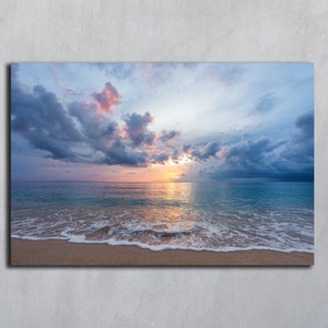 Op de afbeelding: Een landschapsfoto met een zandstrand, schuimige golven en een blauwe zee. De lucht is een mix van blauwe, grijze en roze wolken met een heldere oranje zonsondergang in de verte.