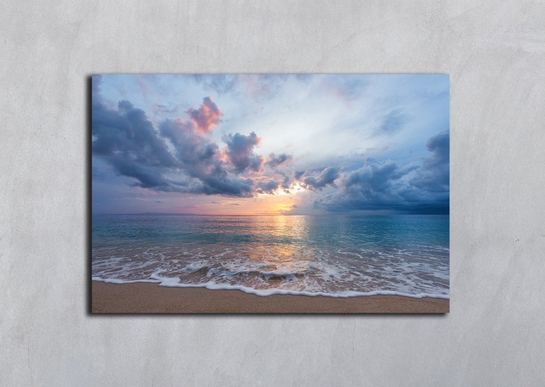 May include: A seascape photograph with a sandy beach, foamy waves, and a blue ocean. The sky is a mix of blue, gray, and pink clouds with a bright orange sunset in the distance.