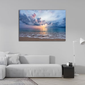 May include: A photograph of a beach scene with a cloudy sky and a sunset over the ocean. The waves are crashing on the shore and the sand is light brown.