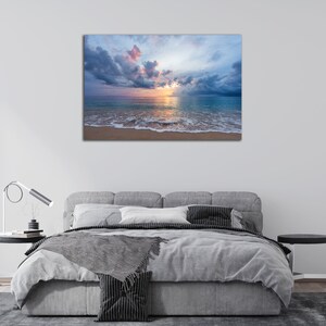May include: A photograph of a beach scene with a cloudy sky and a calm ocean. The sun is setting behind the clouds, casting a warm glow on the water. The sand is white and the waves are gently lapping at the shore.