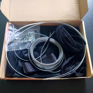 May include: A cardboard box filled with various materials for a DIY project. The box contains black fabric, black ribbon, silver cord, and a roll of black tape.