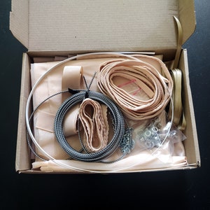 Op de afbeelding: Een open kartonnen doos gevuld met knutselspullen, waaronder een spoel zwart-wit garen, een rol beige lint, een rol beige stof en een zakje zilveren metalen sluitingen.