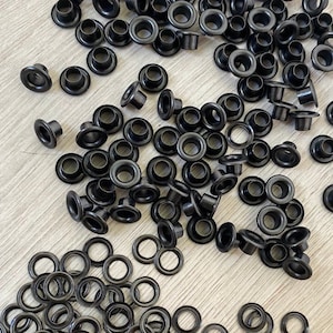 Op de afbeelding: Een grote hoeveelheid zwarte metalen oogjes en ringetjes. De oogjes liggen in een stapel, terwijl de ringetjes verspreid liggen op een licht houten oppervlak.