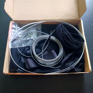 May include: A box filled with black fabric, black ribbon, silver cord, and silver metal rings. The box also contains a clear plastic bag with silver metal clips.