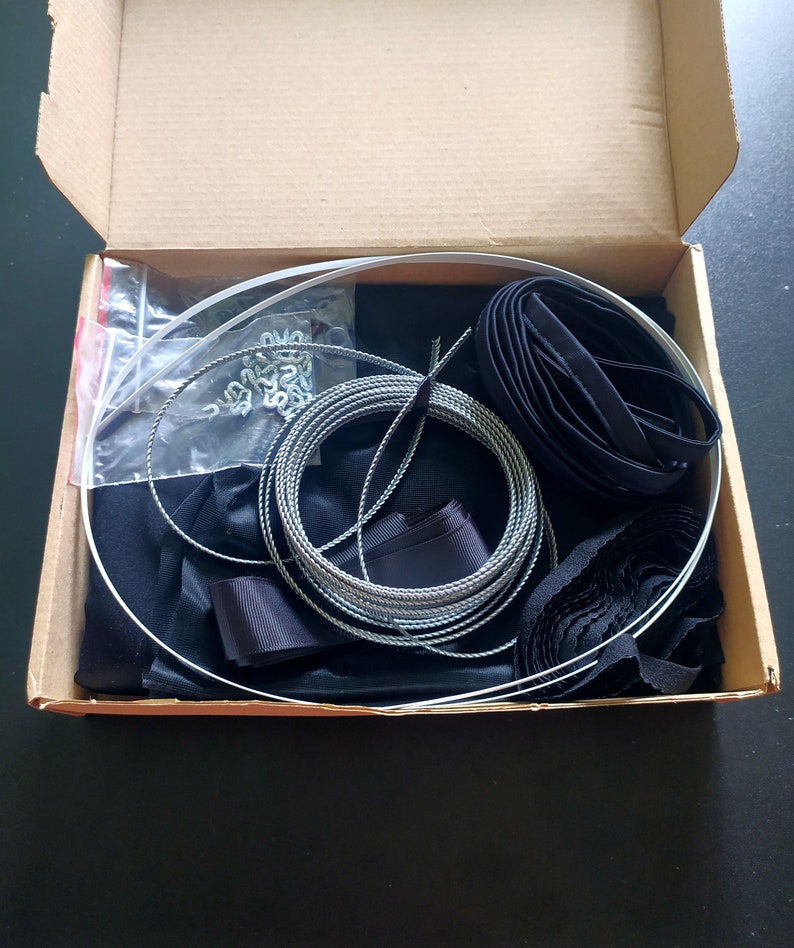 May include: A box filled with black fabric, black ribbon, silver cord, and silver metal rings. The box also contains a clear plastic bag with silver metal clips.