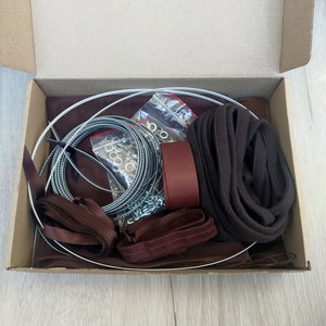 May include: A brown cardboard box containing a selection of craft supplies, including brown ribbon, brown fabric, a metal ring, and a bag of metal eyelets.