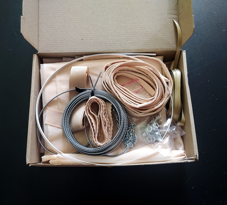 Op de afbeelding: Een open kartonnen doos gevuld met knutselspullen, waaronder een spoel zwart-wit garen, een rol beige lint, een rol beige stof en een zakje zilveren metalen sluitingen.