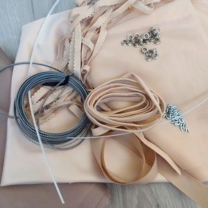 Op de afbeelding: Een verzameling naaigerei, waaronder beige stof, lint, trim, koord, oogjes en metalen bevestigingen.