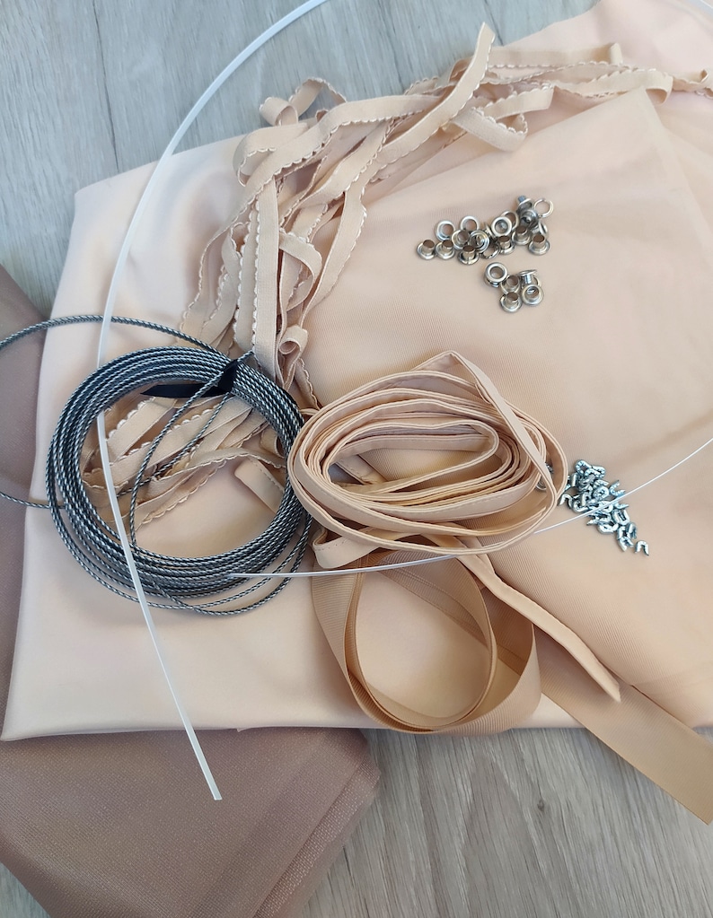 Op de afbeelding: Een verzameling naaigerei, waaronder beige stof, lint, trim, koord, oogjes en metalen bevestigingen.