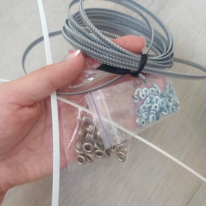 May include: A close-up of a silver metal coil, a bag of silver metal eyelets, and a bag of silver metal hooks. The items are on a light wood surface.