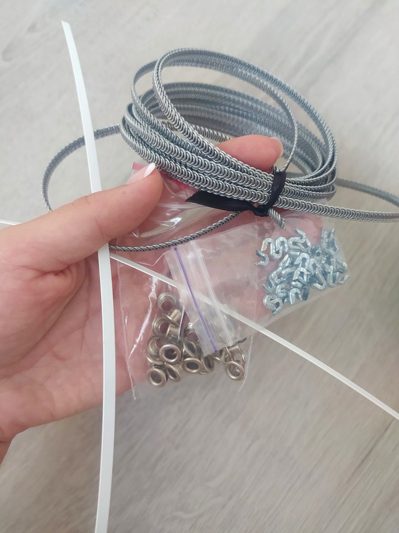 May include: A close-up of a silver metal coil, a bag of silver metal eyelets, and a bag of silver metal hooks. The items are on a light wood surface.