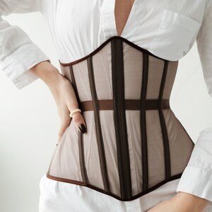 Puede incluir: Un cors&eacute; marr&oacute;n y beige con detalles de cinta marr&oacute;n. El cors&eacute; est&aacute; hecho de una tela transparente y tiene un escote en V.