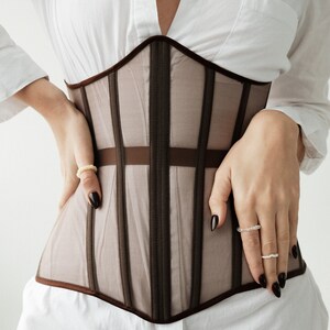 Puede incluir: Un cors&eacute; de malla marr&oacute;n con ribetes de sat&eacute;n negro. El cors&eacute; tiene una estructura de ballenas y est&aacute; dise&ntilde;ado para ajustar la cintura.