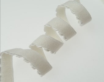 May include: A length of white, scalloped-edge ribbon, spiraling across a white background. The ribbon has a soft, velvety texture and a delicate, decorative edge. The ribbon is likely used for crafting, sewing, or gift wrapping.