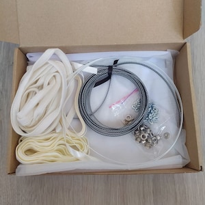 May include: A cardboard box filled with supplies for a DIY project. The box contains white fabric ribbon, a clear plastic circle, a roll of silver wire, and a bag of metal eyelets.