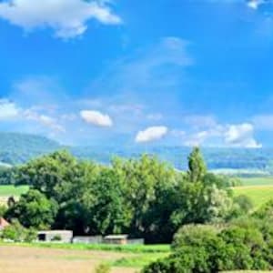 Könnte beinhalten: Panoramablick auf eine Landschaft unter einem hellblauen Himmel mit weißen Wolken. Im Vordergrund grüne Felder und Bäume, mit einer Bergkette in der Ferne. Eine ruhige, natürliche Szene.