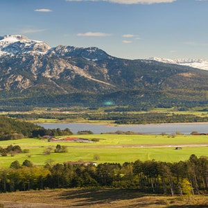 Può includere: Vista panoramica di una catena montuosa con cime innevate sotto un cielo azzurro brillante. Lussureggianti campi verdi e un lago sono visibili nella valle, con alberi e foreste.