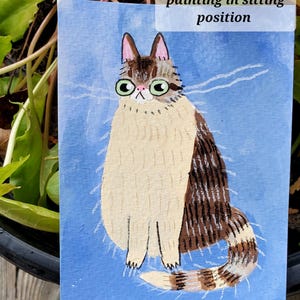 Pode incluir: Uma pintura em aquarela de um gato sentado. O gato tem pelo marrom e branco, &oacute;culos verdes e uma cauda listrada. O fundo &eacute; azul claro. O texto na imagem diz "one cat mini painting in sitting position."