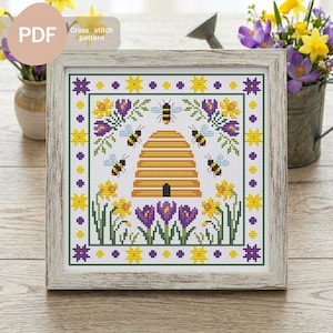 May include: A framed cross-stitch pattern featuring a beehive, bees, and flowers in yellow, purple, and green. The text "PDF Cross stitch pattern" is in the upper left corner. The frame is a light, weathered wood.