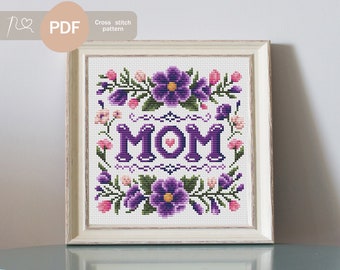 Puede incluir: Patrón de punto de cruz con la palabra "MOM" en morado con un pequeño corazón rosa en el centro. La palabra está rodeada de un borde floral con flores moradas y rosas.