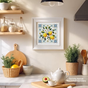 May include: A framed cross-stitch artwork featuring lemons, white blossoms, and green leaves. The artwork is set against a blue and yellow geometric pattern. The kitchen scene includes a teapot, teacup, and a basket with a lemon.