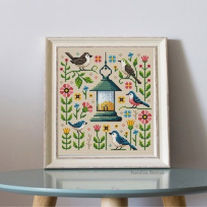 May include: Cross-stitch embroidery of a birdhouse with birds and flowers. The birdhouse is green with a yellow roof and is surrounded by blue, pink, and yellow flowers. The birds are blue, green, and brown.