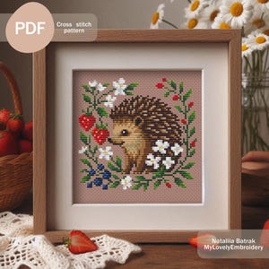 May include: A framed cross-stitch pattern featuring a hedgehog surrounded by flowers, strawberries, and blueberries. The artwork is in a wooden frame with a white mat. The text "PDF" and "Cross stitch pattern" are in the upper left corner.
