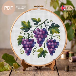 Puede incluir: Un patrón de punto de cruz de uvas moradas y hojas verdes, exhibido en un bastidor de bordado de madera. El diseño está sobre un fondo blanco e incluye el texto "Cross stitch pattern" y "PDF".