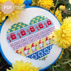 May include: A heart-shaped cross stitch pattern in a blue embroidery hoop featuring horizontal bands of yellow hearts, red padlocks and keys, blue leaves, and three gnomes with red hats on a yellow background, finished with a blue geometric border.