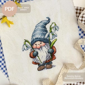 Könnte beinhalten: Kreuzstichmuster eines Zwergs, der einen Strauß weißer Schneeglöckchen hält. Der Zwerg trägt einen blauen Hut mit weißen Streifen, eine braune Jacke und braune Stiefel. Der Hintergrund ist weiß.
