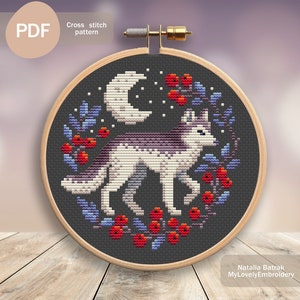 May include: Cross stitch pattern featuring a wolf standing in a wreath of blue and purple flowers with red berries, a crescent moon, and white stars on a dark gray background. The pattern is in a wooden embroidery hoop.
