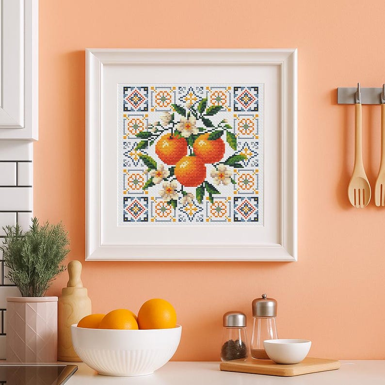 Puede incluir: Una obra de arte en punto de cruz enmarcada que presenta tres naranjas con hojas verdes y flores blancas. Las naranjas est&aacute;n sobre un fondo estampado de azulejos azules, naranjas y blancos. La obra est&aacute; en un marco blanco.