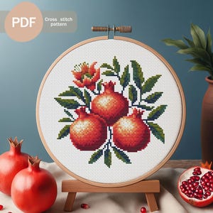 Puede incluir: Un patrón de punto de cruz con tres granadas rojas con hojas verdes y flores rojas, exhibido en un bastidor de bordado de madera. El texto "PDF" y "Cross stitch pattern" son visibles.